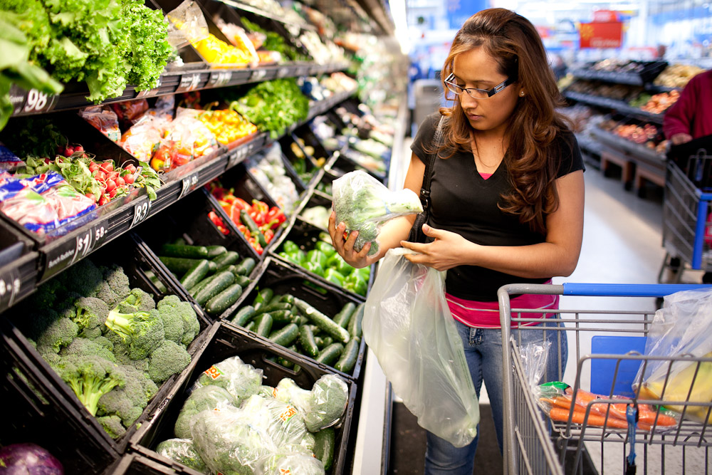 A woman wearing glasses shops for vegetables in a grocery store, placing broccoli into a plastic bag. She stands beside a shopping cart with produce while surrounded by fresh vegetables on display.