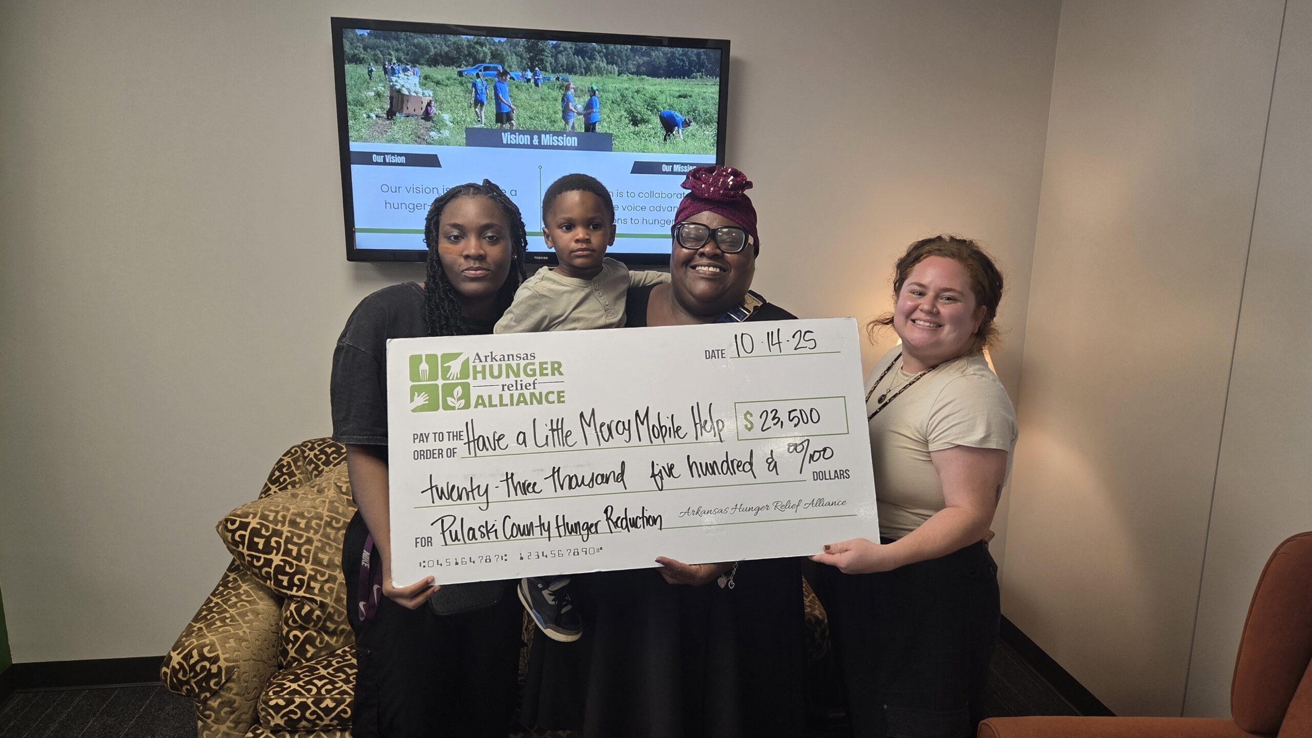 Four people smile while holding a large ceremonial check for $23,500 from Arkansas Hunger Relief Alliance to Make a Life Mason Made, Kids, dated 10-14-23, for Pulaski County Hunger Reduction. A photo and mission statement are in the background.