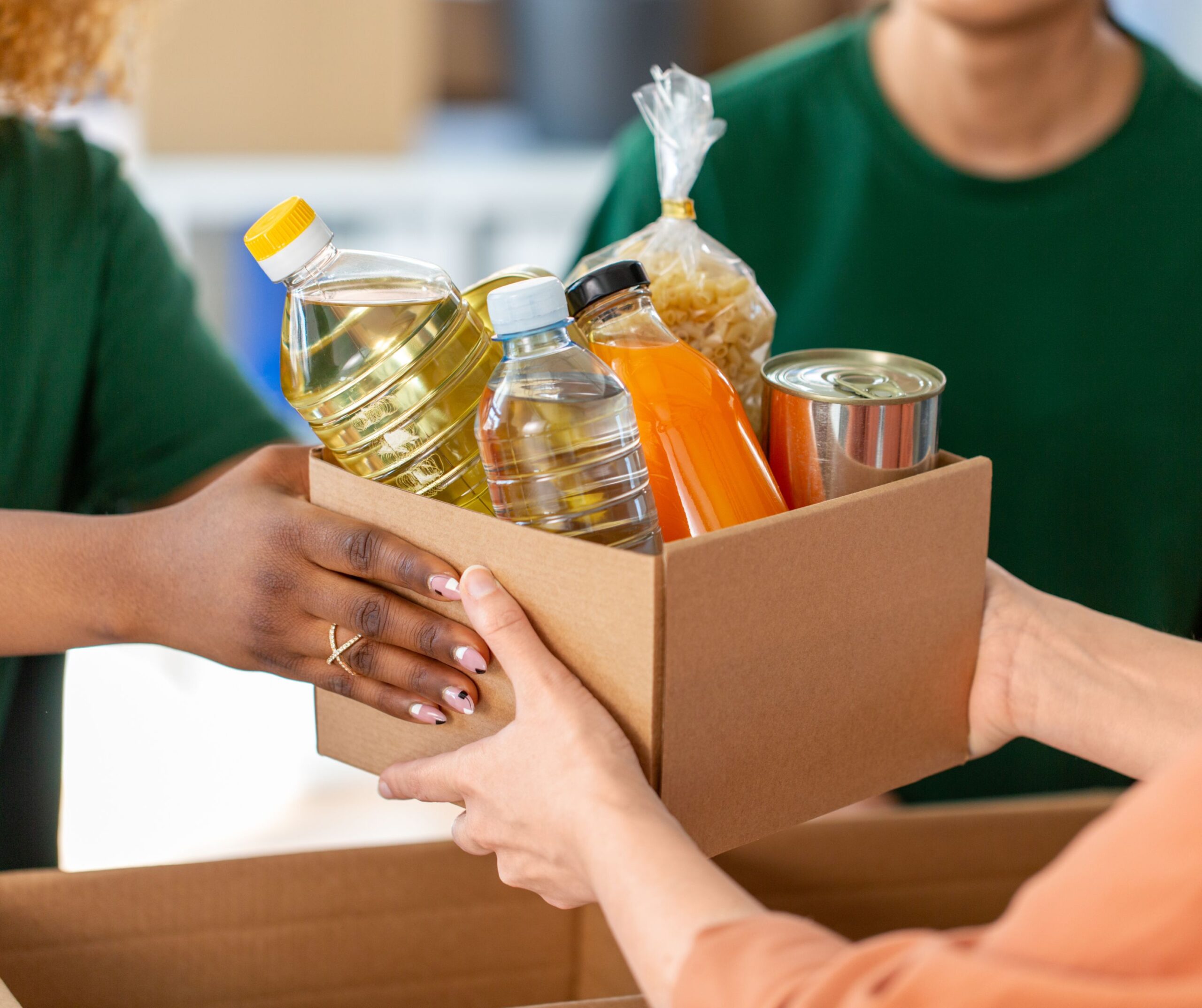 Three people hold a cardboard box filled with food items, including pasta, canned food, a water bottle, oil, and juice. One person hands the box to another, suggesting a food donation or distribution.