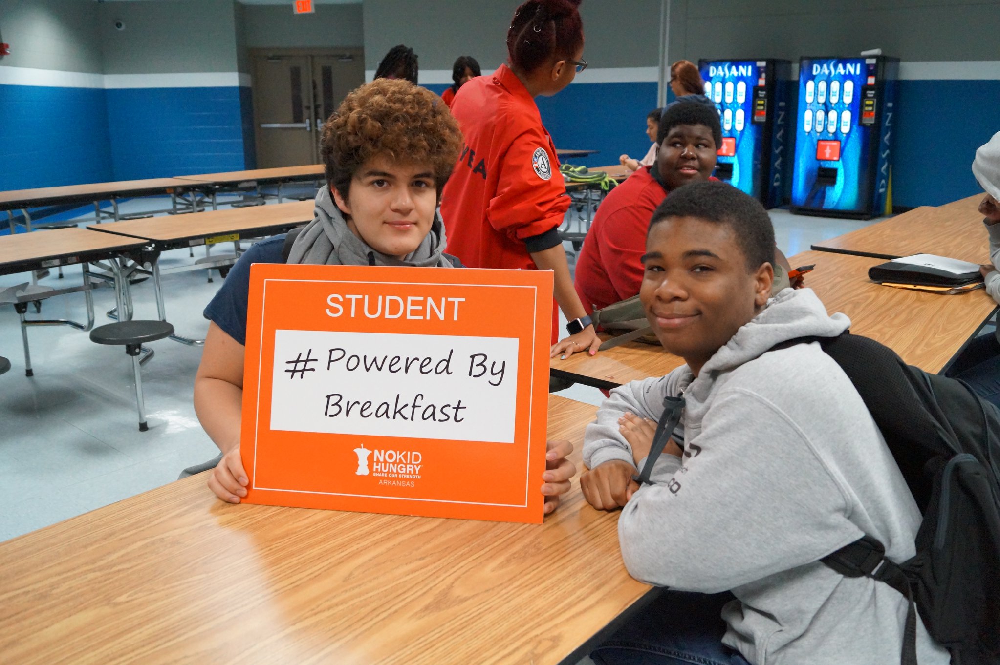 Two students sit at a cafeteria table. One holds an orange sign that says STUDENT #PoweredByBreakfast with the No Kid Hungry logo. Other students and vending machines are visible in the background.