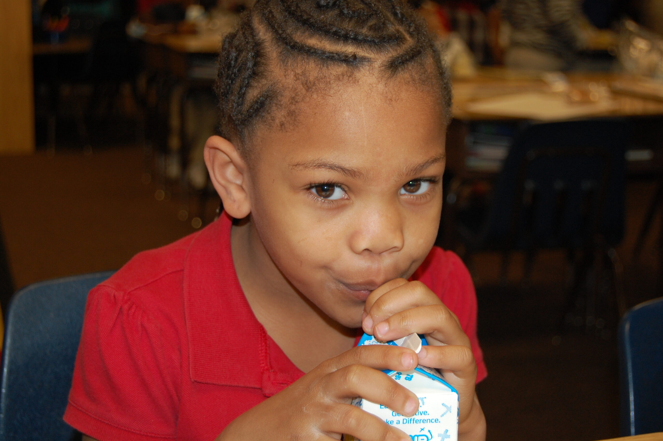 A young child in a red shirt with braided hair sits at a table, looking at the camera while drinking from a small milk carton using a straw. The background shows a classroom setting with chairs and tables.