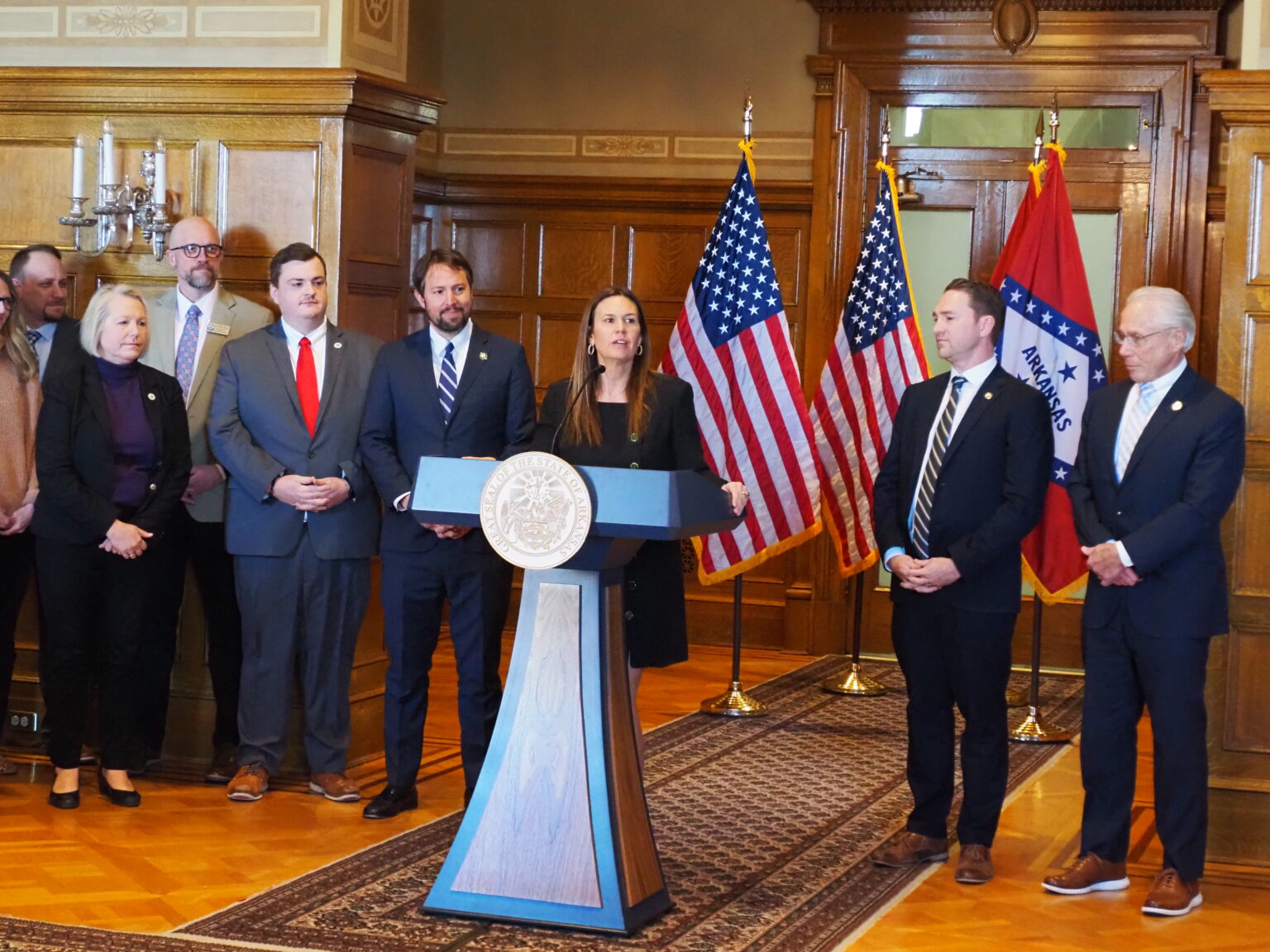 A woman stands at a podium speaking, surrounded by six men and one woman. American and Arkansas state flags are in the background in a wood-paneled room. The group appears to be at a formal press conference or announcement.