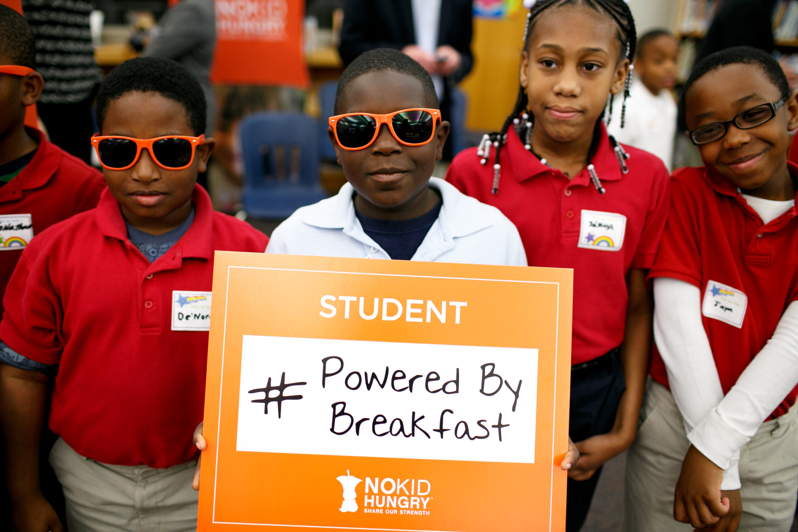 Four children in school uniforms, one holding a sign that says Student #Powered By Breakfast from No Kid Hungry. Two kids wear orange sunglasses. They are standing closely together and smiling in a classroom.