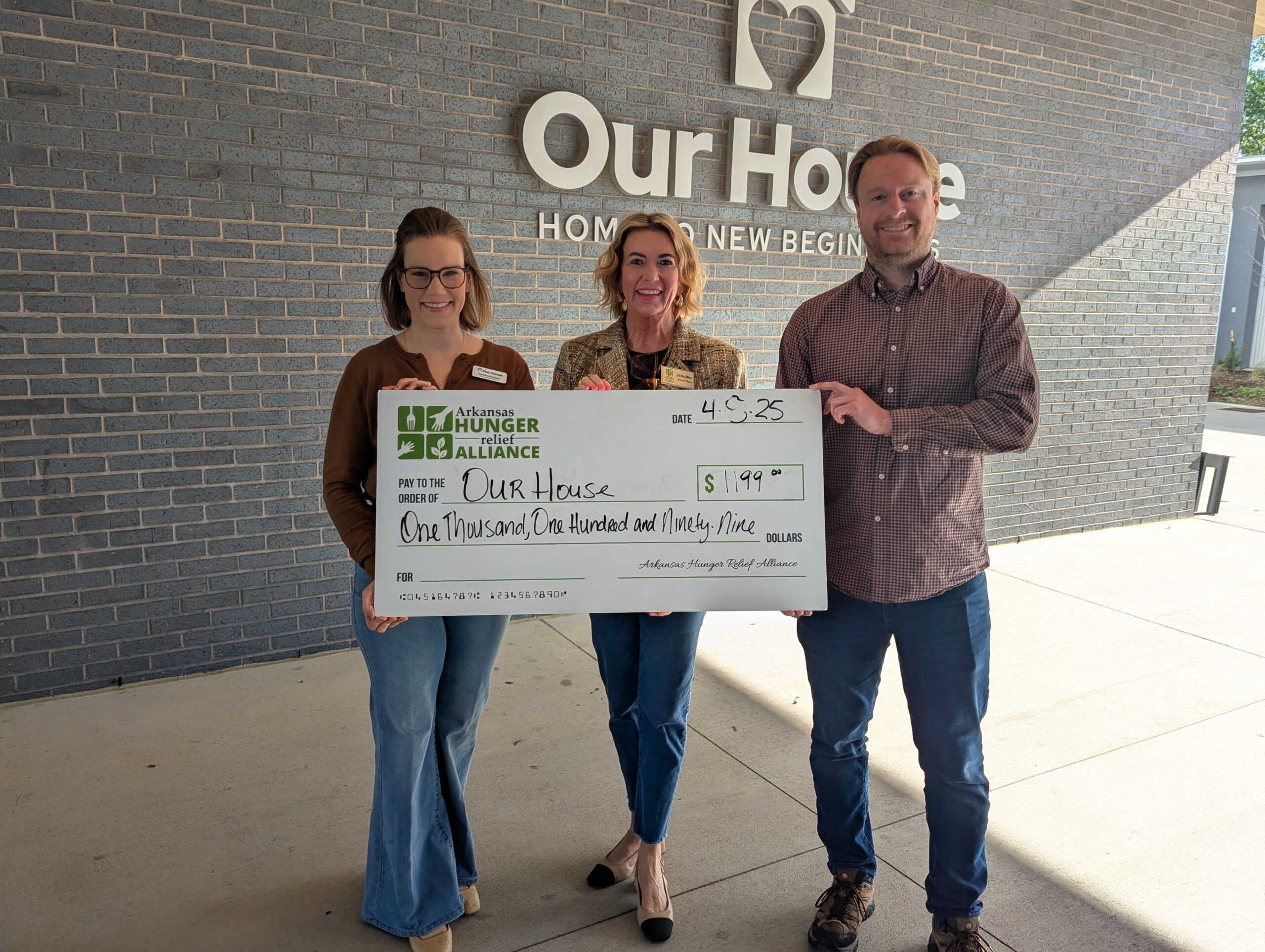 Three people stand in front of a brick wall with an Our House sign, holding a large check for $1,193 from the Arkansas Hunger Relief Alliance, dated 4/25, made out to Our House.