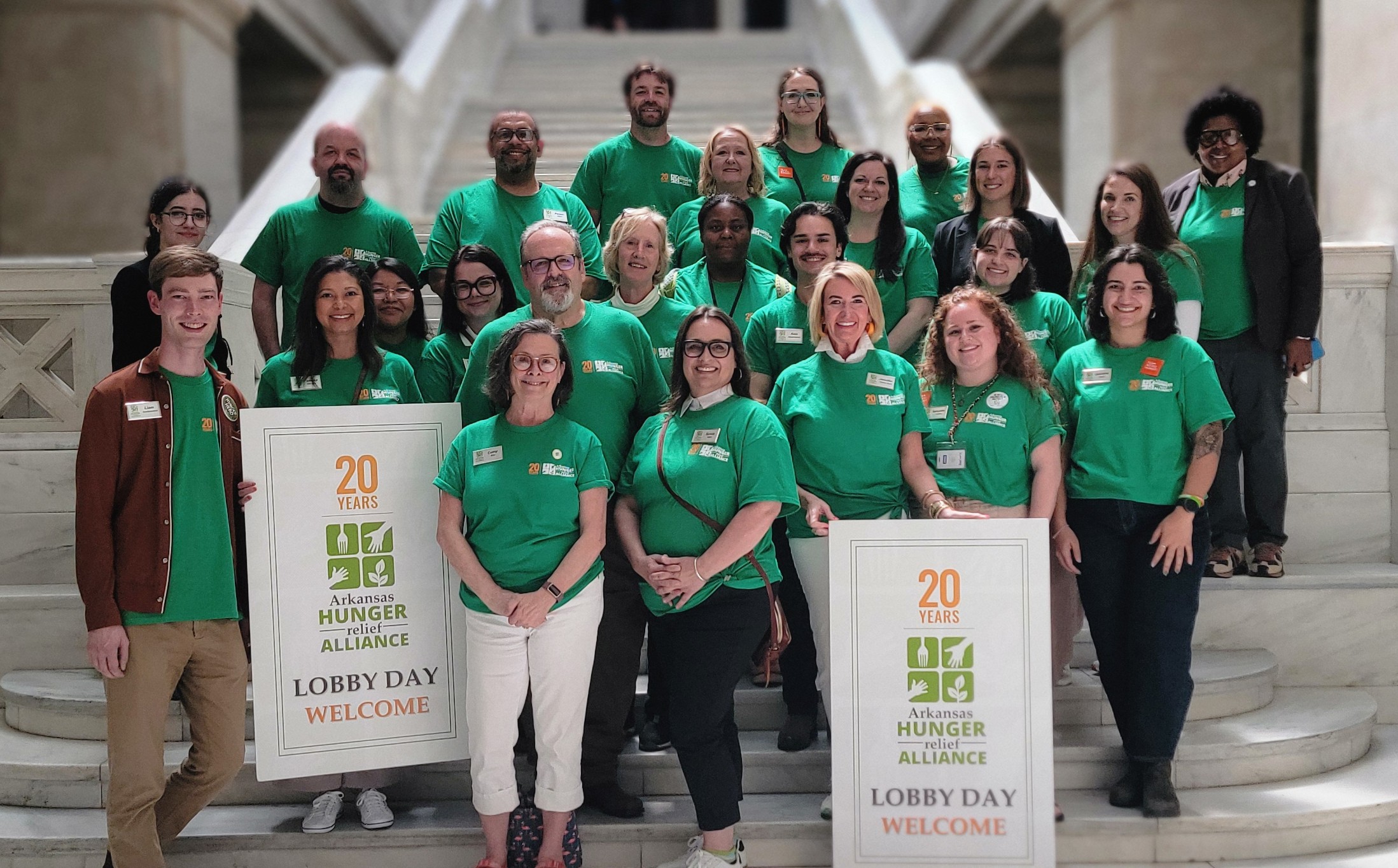 A group of people in matching green T-shirts stand together on marble steps, smiling at the camera. Two people in front hold signs that read “Arkansas Hunger Relief Alliance Lobby Day Welcome.”.