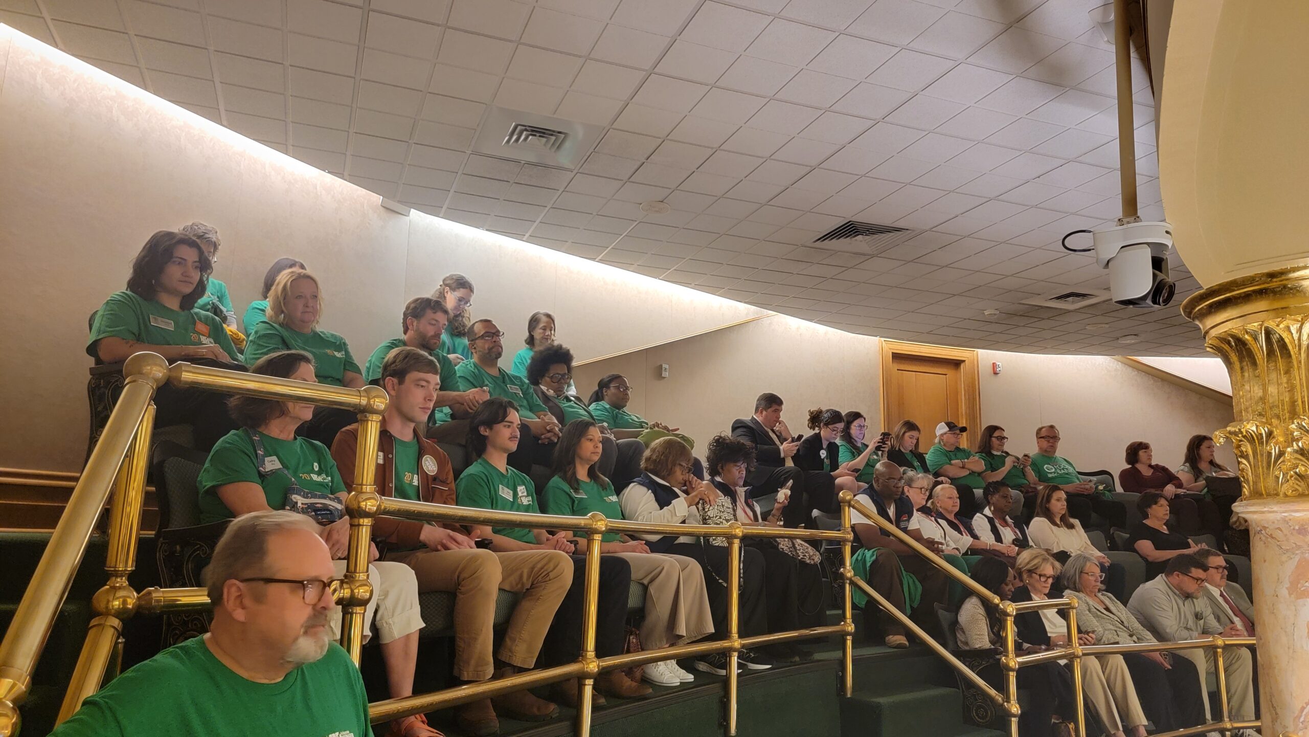 A group of people, many wearing green shirts, sit in stadium-style seating in a balcony area, attentively watching something out of view. Brass railings and a high ceiling are visible in the room.