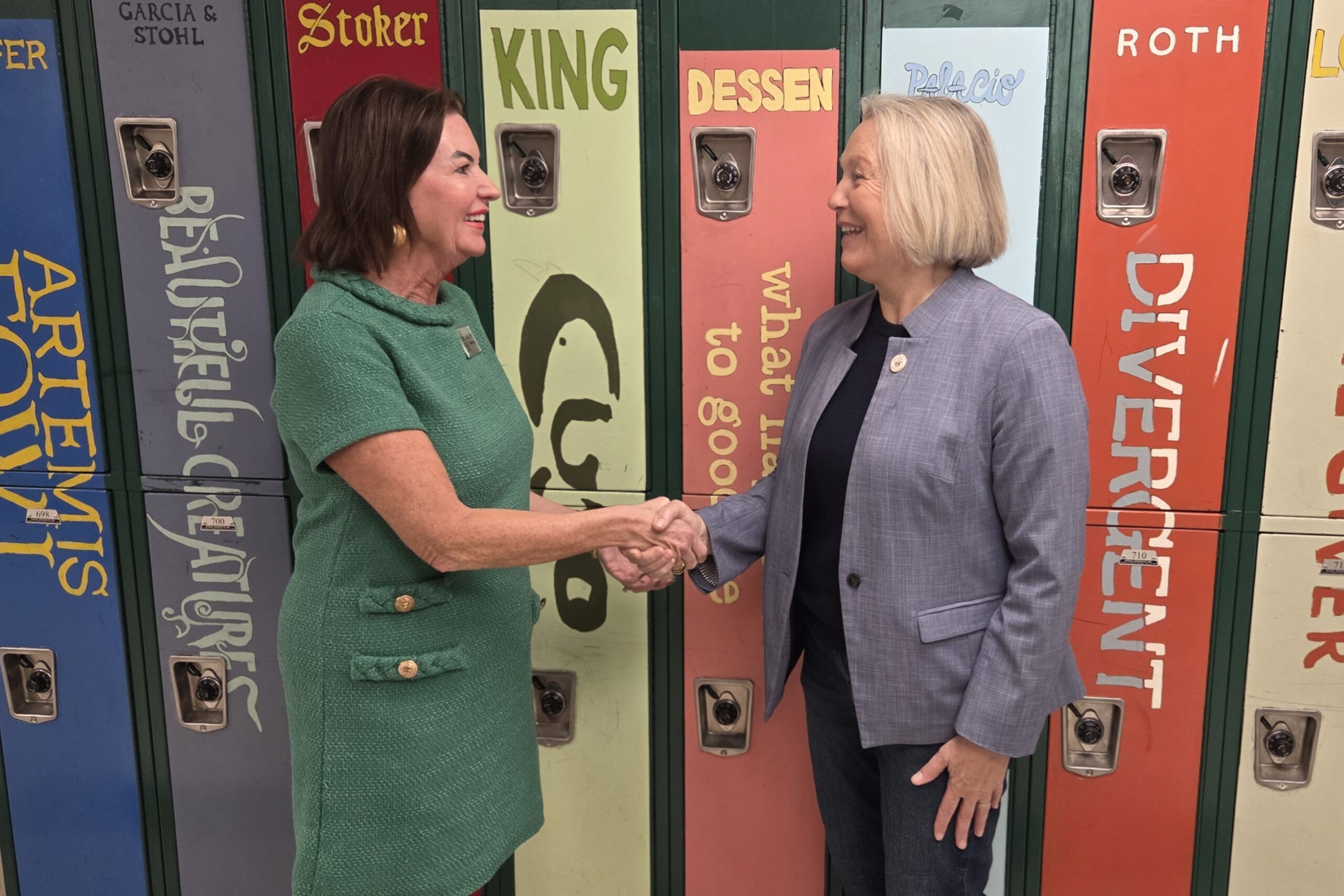 Two women smile and shake hands in front of colorful lockers decorated with popular book titles and authors, including Beautiful Creatures, Divergent, and King.