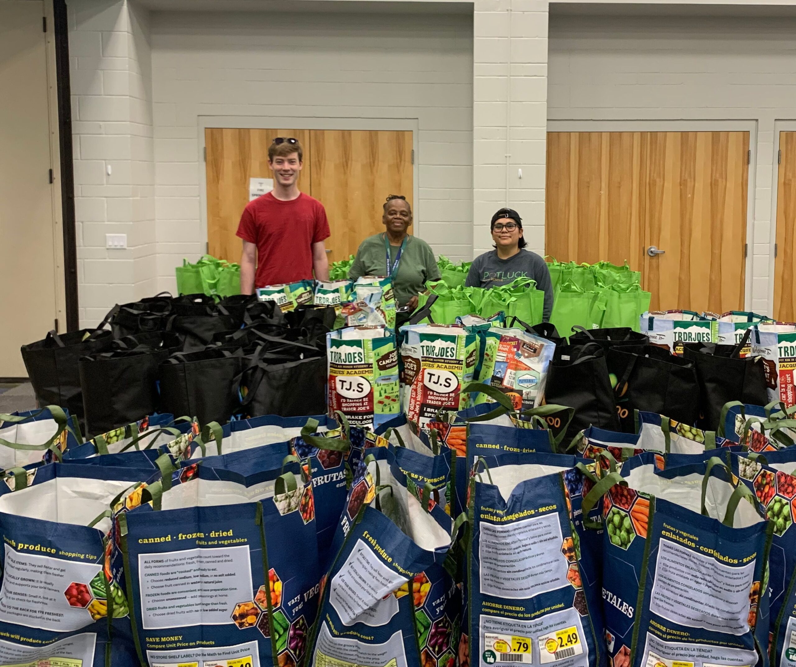 Three people stand behind rows of reusable shopping bags filled with groceries and food items, set up indoors against a wall with wooden doors. The bags appear ready for distribution.