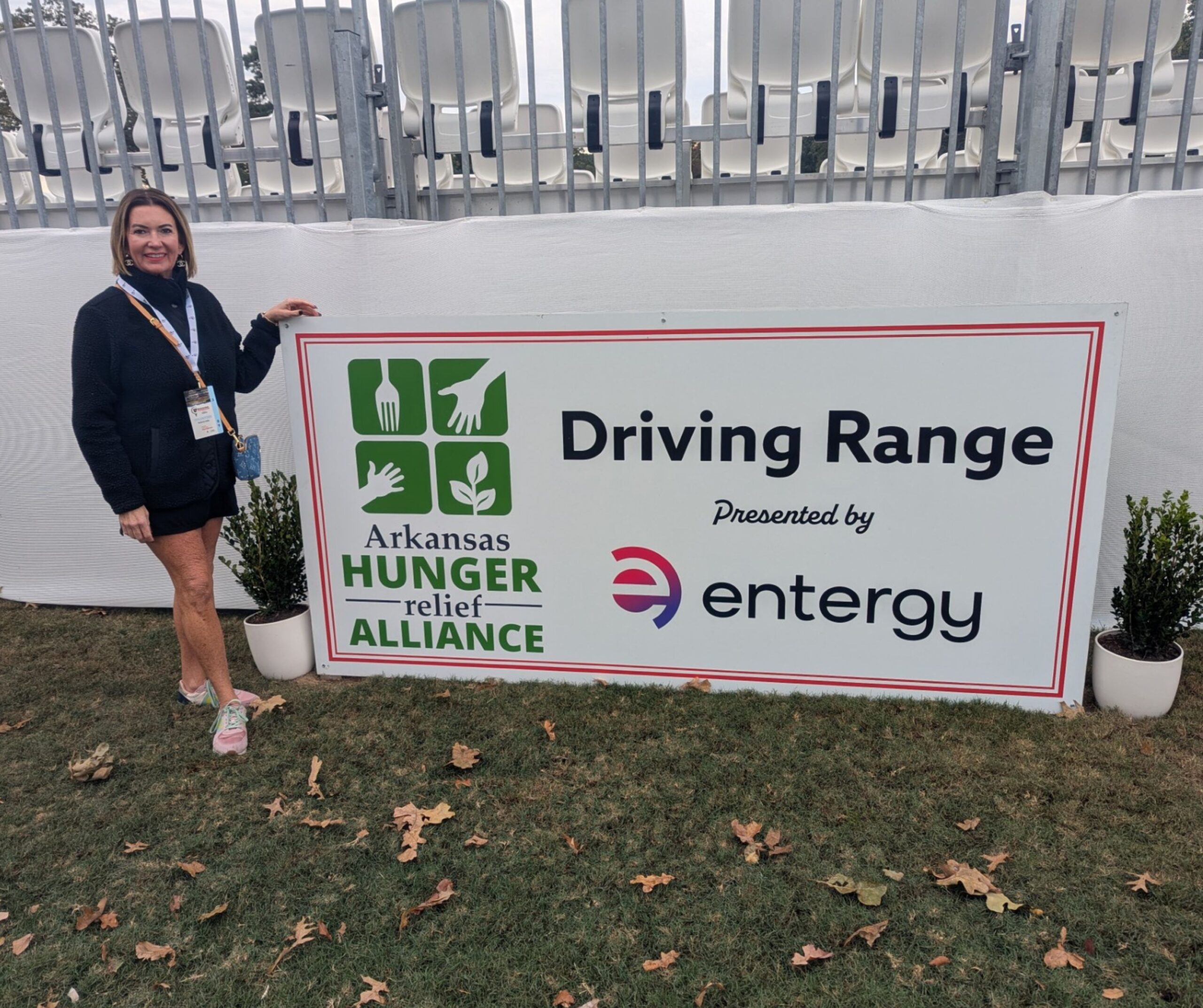 A woman stands next to a large sign that reads “Arkansas Hunger Relief Alliance Driving Range presented by Entergy,” with icons of food and hands. The sign is outdoors on grass, with seating in the background.