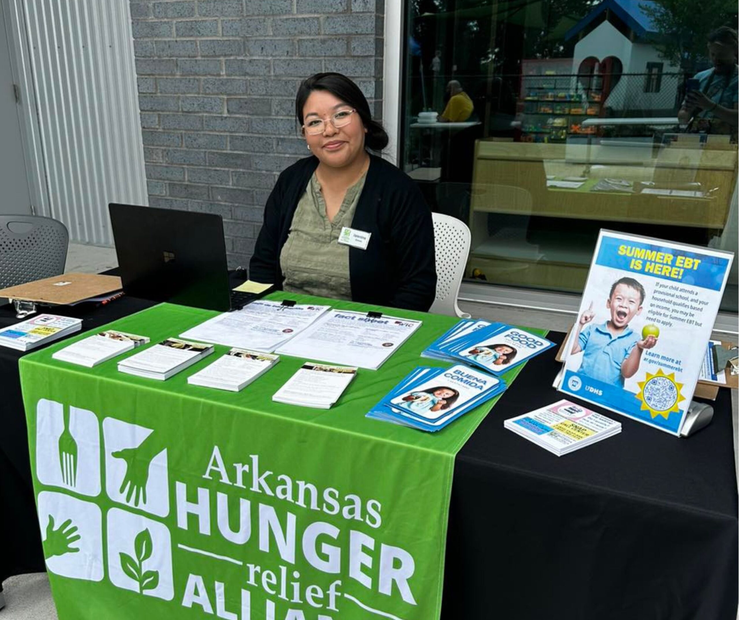 A woman sits at a table covered with an Arkansas Hunger Relief Alliance tablecloth, displaying brochures and a sign about Summer EBT. She is smiling and wearing a name tag, with a laptop in front of her.