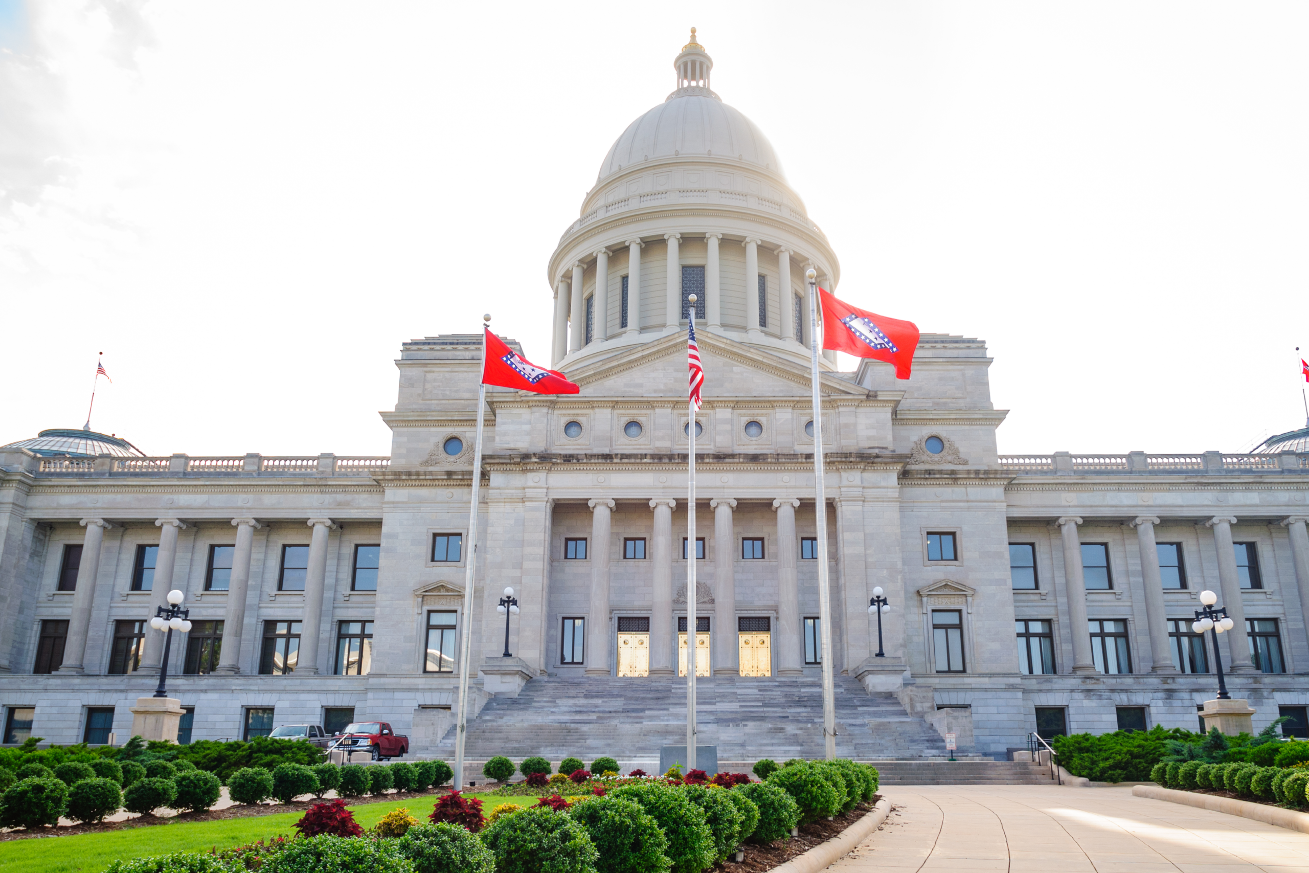 A grand white capitol building with a central dome stands under a clear blue sky, surrounded by lush gardens filled with vibrant, colorful flowers in the foreground.