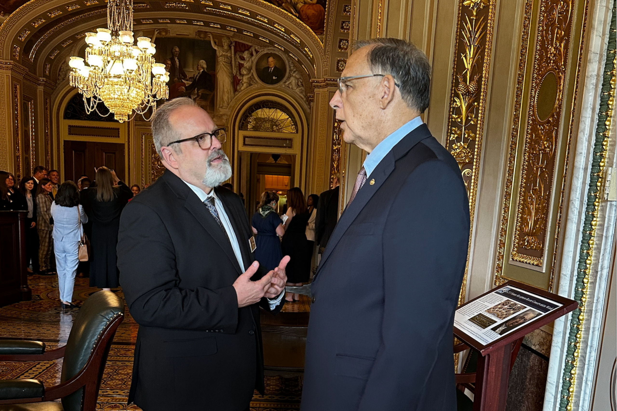 Two men in suits are having a conversation in an ornate room with gold-patterned walls, chandeliers, and several people in the background. One man gestures with his hands while speaking to the other.