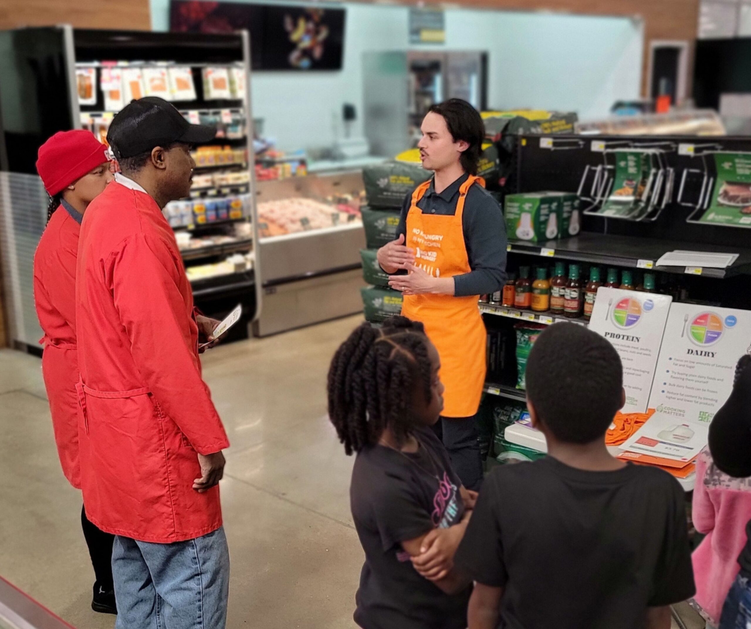 A store employee in an orange apron talks to a group of adults and children near a grocery display with sauces and nutrition charts. The group listens attentively in the store aisle.