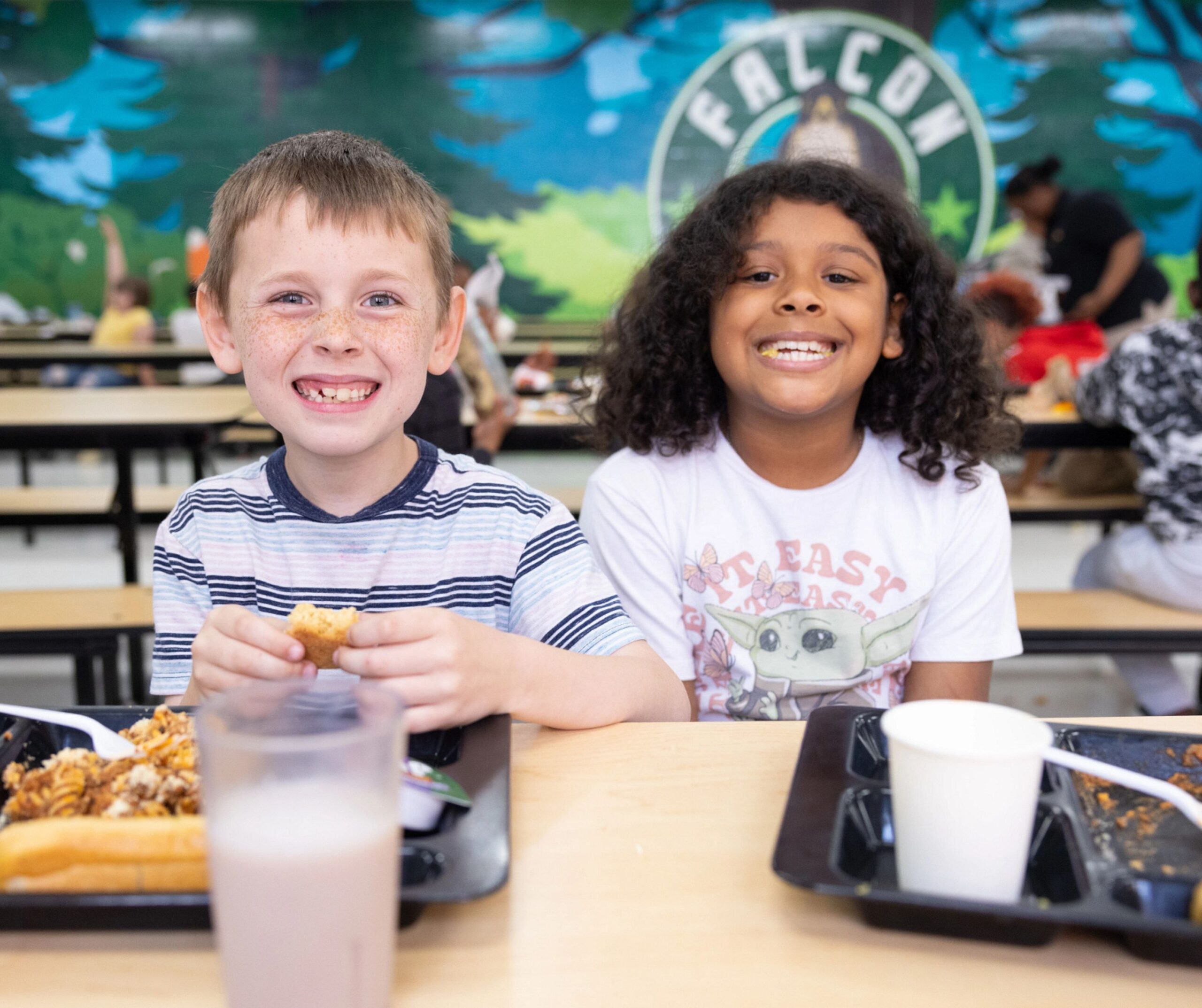 Two children sit side by side at a cafeteria table, smiling at the camera. Food trays, cups, and a colorful mural with the word FALCON are visible in the background.
