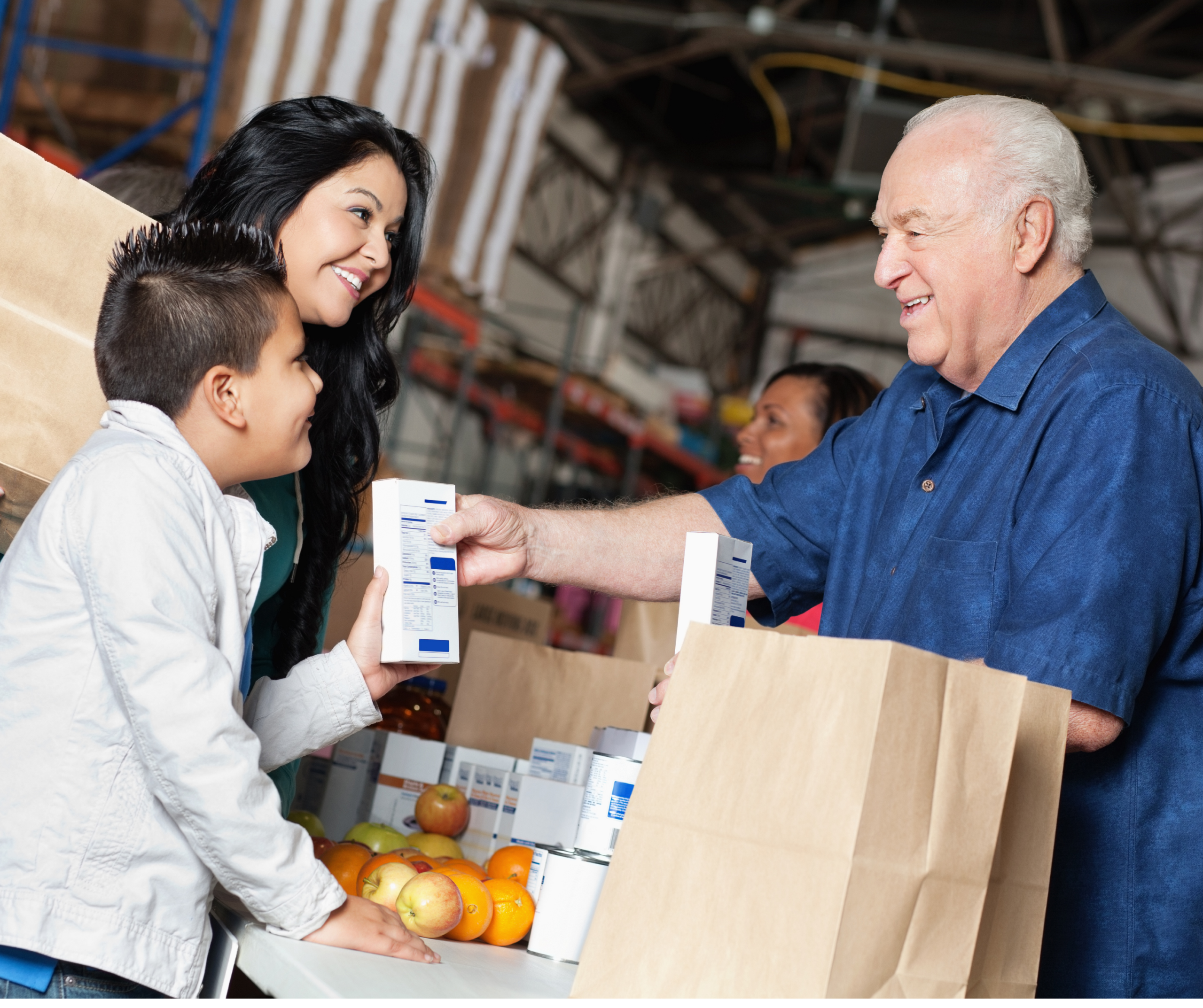 A smiling elderly man hands a food item to a young boy and his mother at a food distribution center. Paper bags filled with groceries and fruit are on the table.