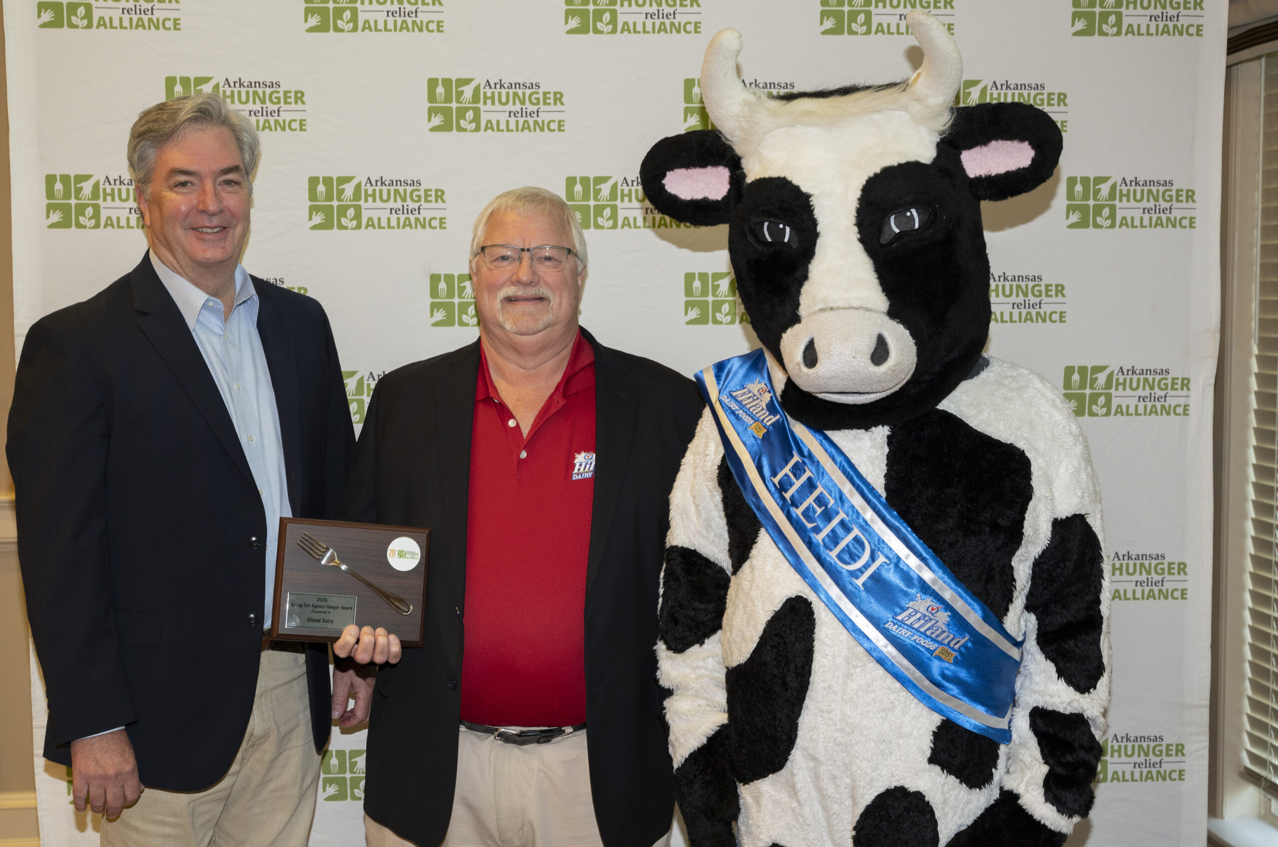 Three men, one in a cow costume with a blue sash reading HEDDY, pose smiling in front of an Arkansas Hunger Relief Alliance backdrop. One man holds a plaque.